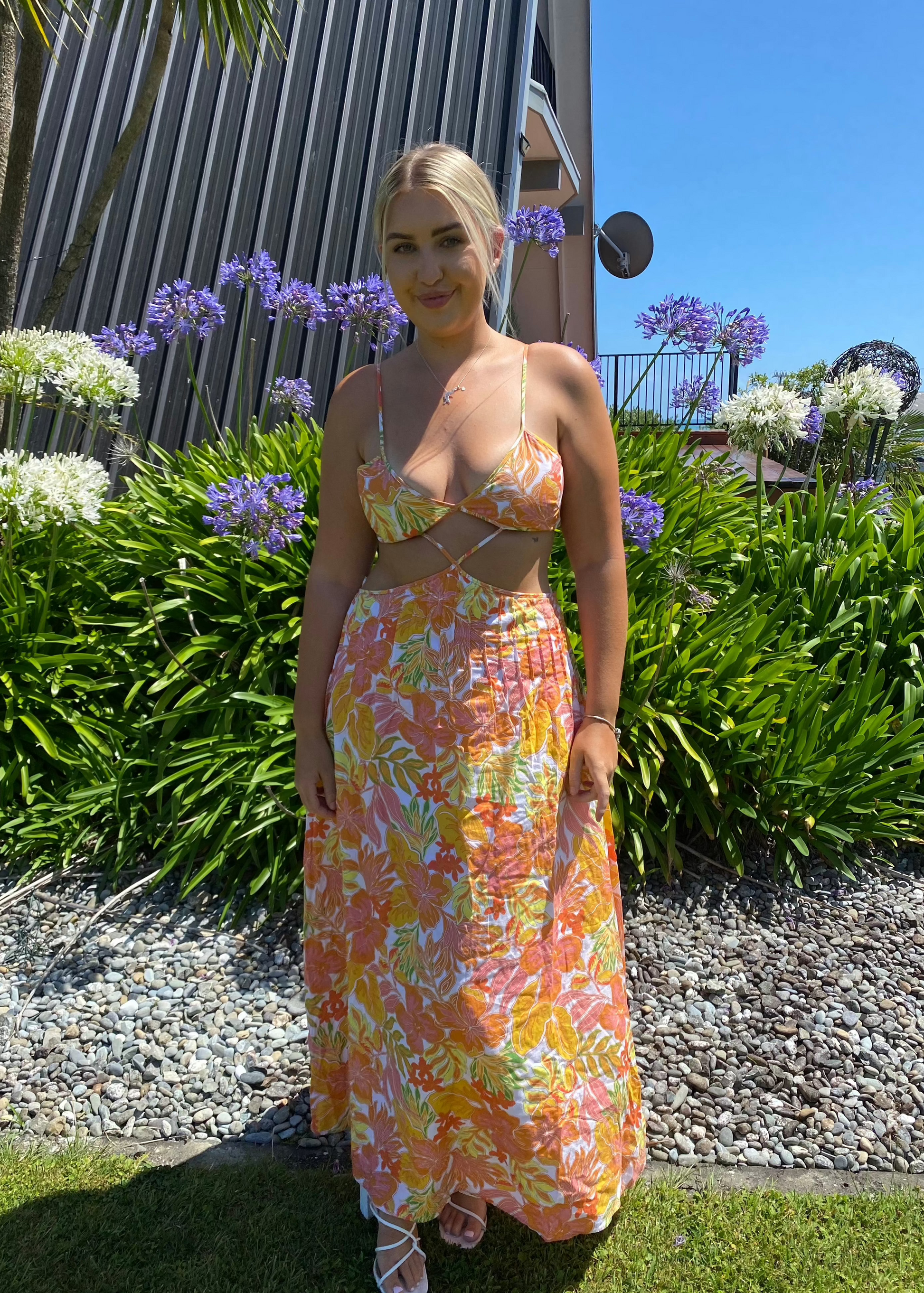 Issy Citrus Aloha Maxi in Floral Print with White, Orange, Pink and Yellow with small hints of green, Size 14. Featuring a lace up back bust area with a open back below with zip on skirt area. Front has stomach cut out with two small straps to attach bust and torso section of dress. Perfect for races, wineries, bottomless brunches etc. Front on full length view of dress.